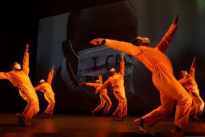 Photo of Woke 2022 performance. Six dancers in orange prison jumpsuits lean backwards in front of a video projection of a man wearing a mask with 
